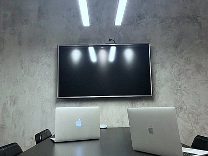 Mesa de reunião vista de frente, com dois notebooks abertos e televisor de apoio.