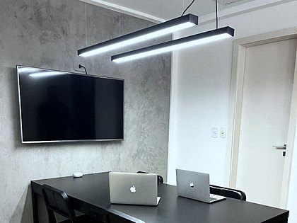 Sala de reunião com mesa preta, televisor na parede de concreto e luminárias lineares.